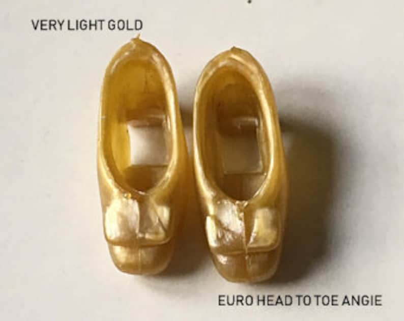 May include: Pair of gold-colored plastic doll shoes with a bow detail. The shoes are labeled "VERY LIGHT GOLD" and "EURO HEAD TO TOE ANGIE".