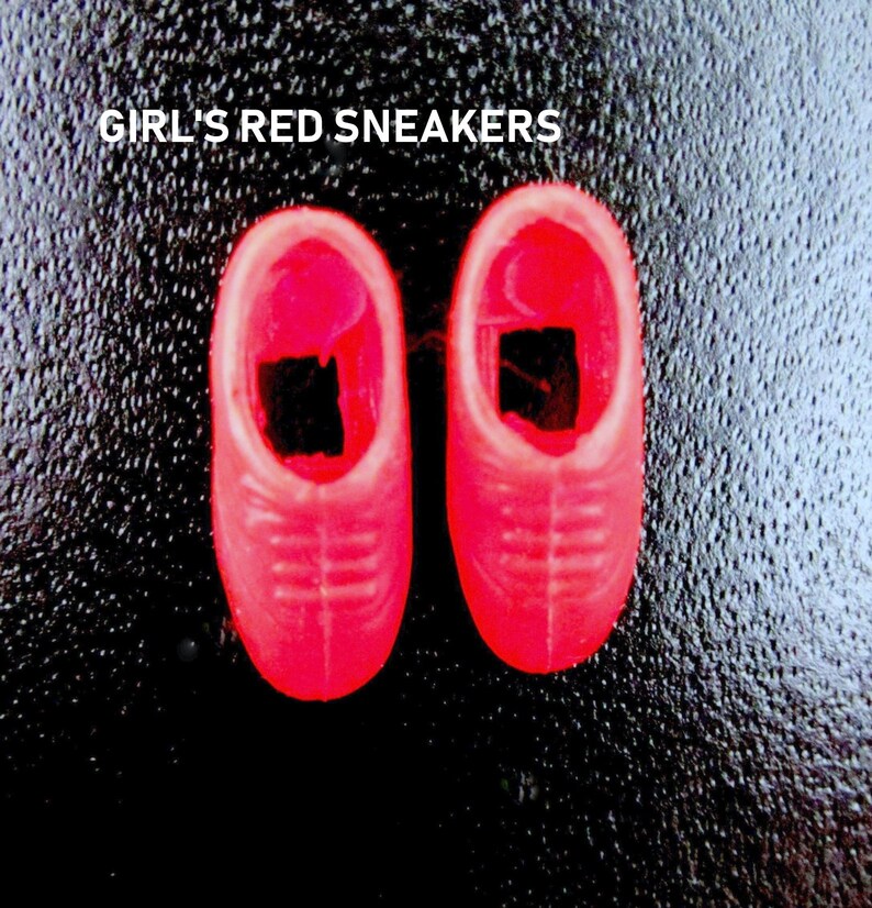May include: A pair of bright red sneakers for a doll. The sneakers have a simple design with white stitching and a black accent on the toe.