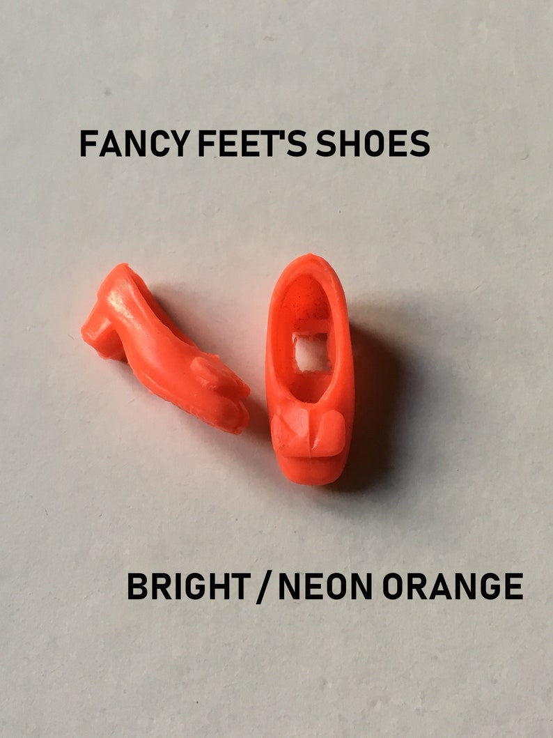 May include: A pair of bright orange plastic doll shoes. "FANCY FEET'S SHOES" and "BRIGHT/NEON ORANGE" are printed on the white background.