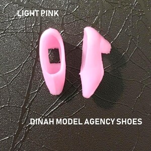 May include: Two light pink plastic doll shoes. The shoes are in the shape of a slipper. The text "DINAH MODEL AGENCY SHOES" is printed below the shoes.