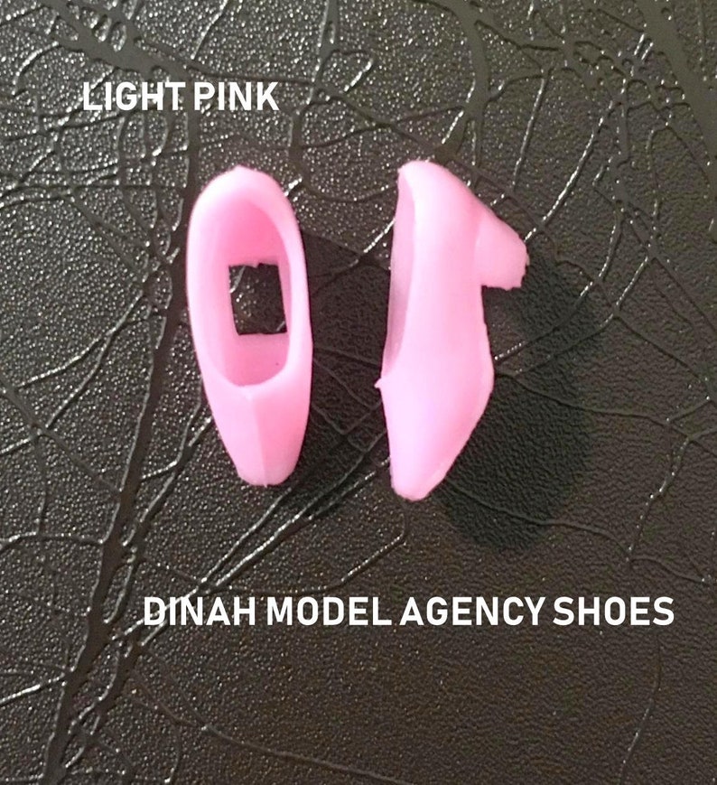 May include: Two light pink plastic doll shoes. The shoes are in the shape of a slipper. The text "DINAH MODEL AGENCY SHOES" is printed below the shoes.