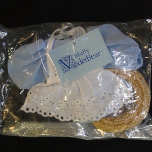 May include: A light blue dress with white lace trim and a straw hat. The dress and hat are packaged together in a clear plastic bag. The bag has a label that reads "Muffy VanderBear".
