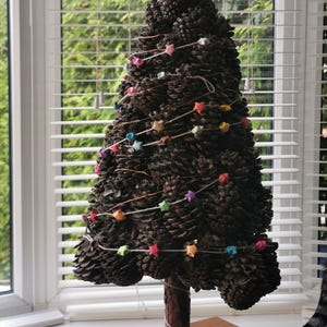 May include: A handcrafted Christmas tree made of pinecones, decorated with colourful star-shaped ornaments strung on delicate wire. The tree stands on a wooden base, creating a rustic and festive holiday decoration. The tree is approximately 30 cm tall.