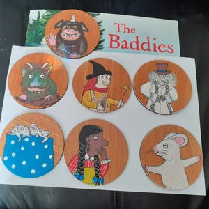 May include: Seven circular wooden coasters featuring colourful illustrations of characters and scenes from the book The Baddies. The designs include a horned monster, a witch with a wand, a ghost in a top hat, mice in a pocket, a person with braids, and a white mouse.