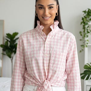 May include: A pink and white gingham button-down shirt with a ruffled collar, tied at the waist. The shirt has long sleeves and a single chest pocket. The person is wearing white jeans.