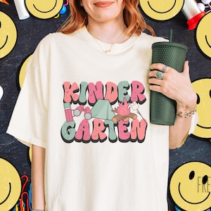 May include: A cream-coloured t-shirt featuring the word "KINDERGARTEN" in a retro font with camping-themed graphics. The design includes a tent, binoculars, and a campfire. A dark green tumbler is held in the hand.