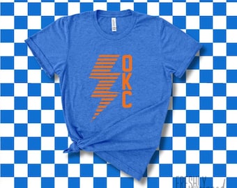 May include: Blue t-shirt with orange OKC lightning bolt graphic. The shirt is a crew neck and has short sleeves. The background is a blue and white checkered pattern.