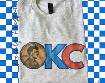 May include: A heather gray t-shirt featuring the letters "OKC" in blue and orange, with a penny graphic depicting a man's profile. The penny graphic includes the text "IN GOD WE TRUST" and "2025". The shirt is on a blue and white checkered background.
