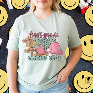 May include: A pale green t-shirt with a colourful graphic. The design includes the words "first grade", camping imagery, and the words "Camping Crew". The shirt is a casual style, suitable for everyday wear.