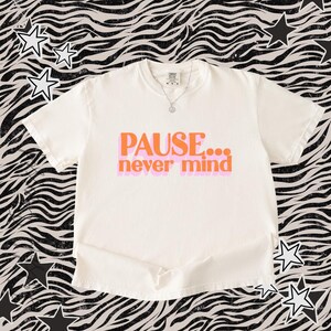May include: An off-white short-sleeve t-shirt featuring the orange text PAUSE... never mind with a light pink shadow effect behind the words. The shirt has a crew neckline and a silver-toned chain necklace with a small circular pendant.