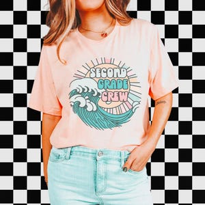 May include: A peach-colored t-shirt featuring a circular graphic with the words "SECOND GRADE CREW" in a stylized font. The graphic includes a wave design in teal and white, with a sunburst and pastel stripes. The shirt is worn by a person.