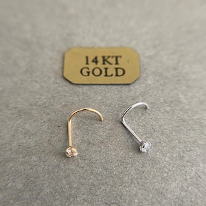 Puede incluir: Dos anillos de nariz con pequeñas piedras transparentes. El anillo de nariz de la izquierda es de oro y el anillo de nariz de la derecha es de plata. La etiqueta dice "14KT GOLD".