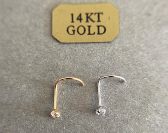 May include: Two 14kt gold nose rings with a small clear stone on the end. The rings are shown on a gray surface with a label that reads "14KT GOLD".