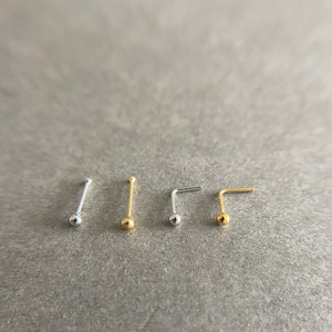 May include: Five nose rings with a ball end. The rings are made of silver and gold metal. The rings are arranged in a row on a gray surface.