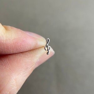 May include: A silver treble clef charm, a musical symbol, is held between two fingers.