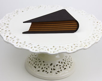 Könnte beinhalten: Ein weißer Kuchenständer mit einem Spitzenmuster, auf dem ein Schokoladenkuchen in Form eines Buches mit braunen und schwarzen Streifen liegt.