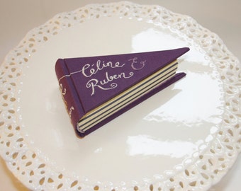 Könnte beinhalten: Ein lilafarbenes Buch mit weißer Beschriftung, auf dem "Celine & Ruben" steht, auf einem weißen Spitzendeckchen.