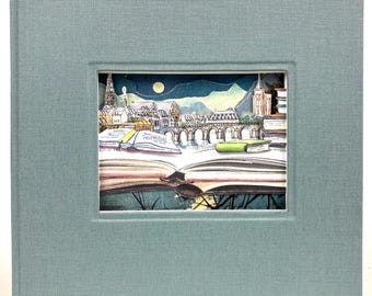 Könnte beinhalten: Ein hellblaues Buch mit einem quadratischen Ausschnitt, der eine 3D-Papierkunstszene zeigt. Die Szene zeigt eine europäische Stadt mit einer Brücke, Gebäuden und einem Vollmond. Offene Bücher und Bücherstapel sind ebenfalls zu sehen.