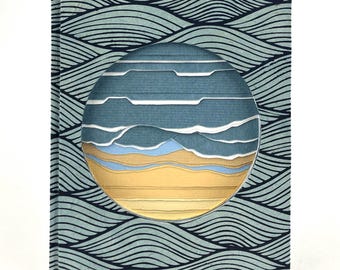 Könnte beinhalten: Ein Hardcover-Buch mit einem blau-weißen Wellenmuster. Ein kreisförmiger Ausschnitt zeigt eine mehrschichtige Papierkunstszene eines Strandes mit blauem Wasser und goldenem Sand. Der Einband des Buches ist helltürkis.