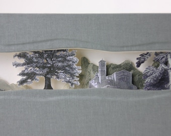 Könnte beinhalten: Ein graues Stoffpanel mit einem Ausschnitt, der einen Papier-Collage eines Landschaftsbildes mit Bäumen, einem Gebäude und Bergen zeigt. Die Collage ist in Grautönen und Schwarz gehalten.