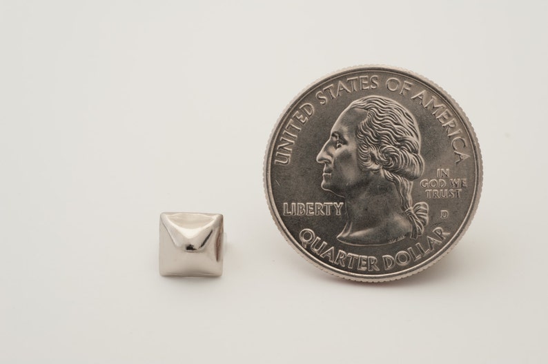 May include: A silver pyramid-shaped metal rivet next to a US quarter dollar coin. The coin features the profile of George Washington and the words "United States of America", "Liberty", "In God We Trust", and "Quarter Dollar".