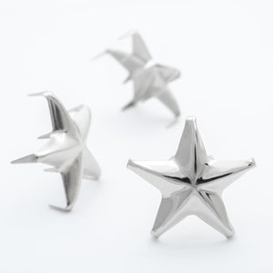 May include: Three silver star-shaped metal embellishments. The stars are three-dimensional and have a pointed tip.