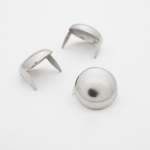 May include: Three silver-colored upholstery tacks with rounded heads and two prongs. The tacks are arranged on a white background, with one tack in the foreground and two in the background. The tacks are made of metal and have a shiny finish.