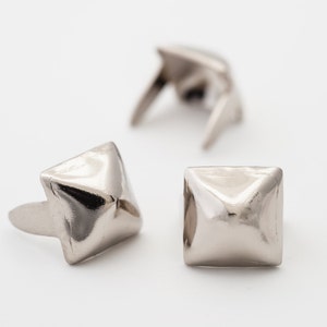 May include: Three silver pyramid-shaped metal studs with a square base. The studs have a shiny finish and are used for decorative purposes.