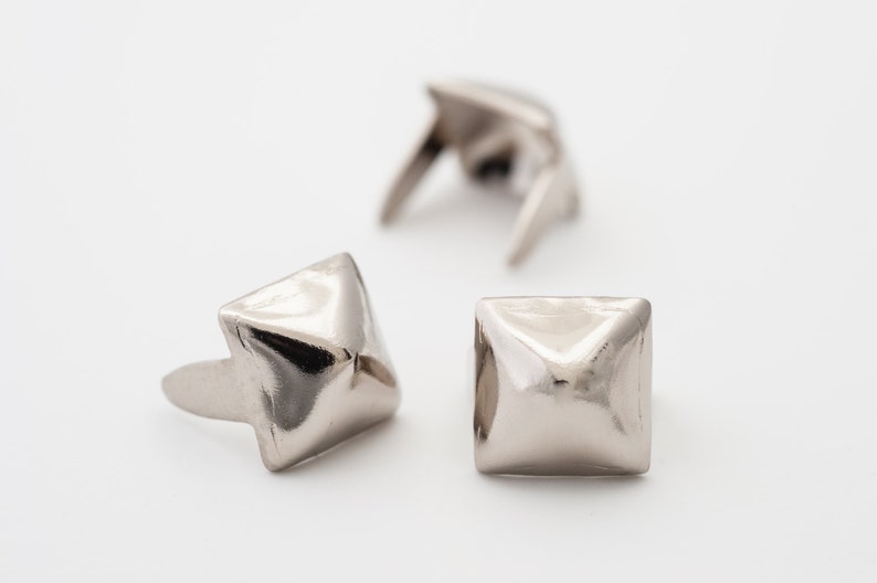 May include: Three silver pyramid-shaped metal studs with a square base. The studs have a shiny finish and are used for decorative purposes.