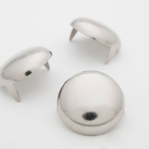 May include: Three silver-colored, dome-shaped metal rivets with short, flat prongs. The rivets are arranged on a white background, with the light reflecting off their smooth, rounded surfaces. These are likely used for leathercraft or other decorative applications.