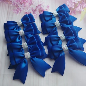 May include: A set of twelve royal blue satin bows with silver glitter accents. The bows are arranged in a cluster on a white wooden surface.