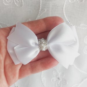 Puede incluir: Un lazo de satén blanco con un centro de perlas y pedrería. El lazo se sostiene en una mano, mostrando su delicado diseño. Este accesorio podría usarse para el cabello o como adorno.