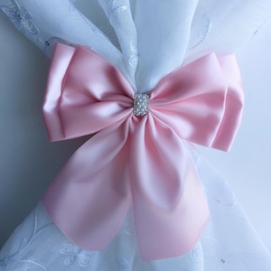 Pode incluir: Um laço grande de cetim rosa claro com um centro retangular adornado com pérolas. O laço é amarrado em volta de uma cortina branca transparente com bordados florais delicados, criando um detalhe decorativo.