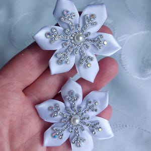 May include: Two white satin flower hair clips. Each clip features a snowflake design with silver rhinestones and a pearl centre. The petals are made of white satin ribbon. The clips are held in a hand.