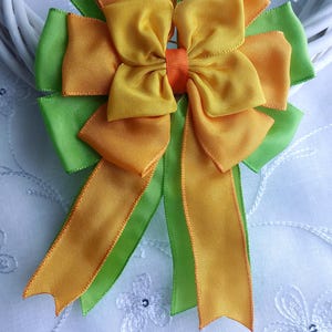 Pode incluir: Um laço decorativo feito de fitas de cetim em camadas em tons de amarelo, laranja e verde. O laço é centralizado e tem caudas longas e fluidas. O fundo é uma superfície branca com bordados florais.