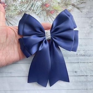 Puede incluir: Un gran lazo de satén azul marino con un centro de purpurina plateada. El lazo está atado con una cola larga.