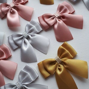 May include: Assortment of satin ribbon bows in gold, silver, and blush pink colors. Each bow has a silver glitter accent in the center.