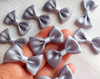Puede incluir: Una colección de lazos de satén plateado. Los lazos son pequeños y delicados, perfectos para agregar un toque de elegancia a las manualidades, los regalos o los accesorios para el cabello.