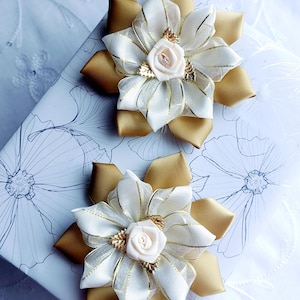 Puede incluir: Dos accesorios florales decorativos para el cabello. Cada uno presenta una base de satén dorado, capas de cintas marfil y un delicado centro de rosa marfil. Acentos dorados y una superposición transparente añaden detalles. Los accesorios descansan sobre una superficie con estampado floral.