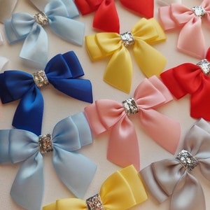 May include: Assortment of satin ribbon bows in various colours, including blue, yellow, pink, red, and grey. Each bow has a silver glitter accent in the centre.