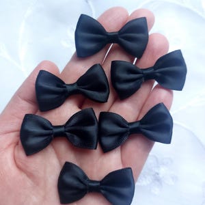 May include: Seven small, black satin bow ties are displayed. Each bow tie is neatly tied, highlighting their symmetrical design. The bows are a solid black colour, and the image is set against a white, textured background.