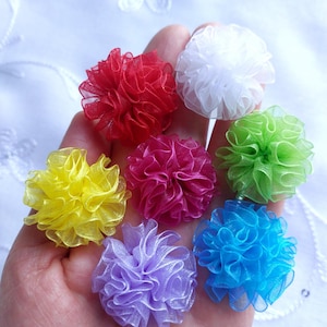 Pode incluir: Uma mão segurando sete pequenas flores de tecido franzido em várias cores: vermelho, branco, verde, amarelo, rosa, roxo e azul. As flores têm uma aparência delicada e arejada, possivelmente feitas de tule.