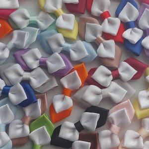 May include: A collection of numerous small hair bows, each featuring a solid-coloured satin ribbon base layered with a smaller white satin ribbon bow on top. The bows appear in a wide variety of colours including red, blue, yellow, green, and pink.