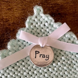 May include: A light green woven fabric with a white ribbon tied in a bow. A silver charm with the word "Pray" is attached to the ribbon.