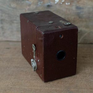 May include: A vintage brown box camera with a textured leather-like exterior. The camera has a lens, a viewfinder, and various metal details. The classic design suggests it's a collectible item, perfect for photography enthusiasts or vintage decor.