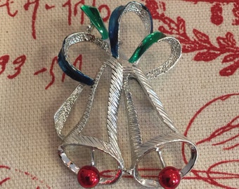 May include: A silver-toned Christmas bell brooch with two bells and a bow. The bow has green and blue enamel accents. Each bell has a small, red, round ornament at the bottom. The brooch is set against a textured, off-white background.