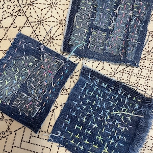 Może przedstawiać: Trzy kwadraty dżinsu z widocznymi detalami szytymi ręcznie. Każdy kwadrat ma inny wzór kolorowych ściegów z nici, w tym różowy, żółty i niebieski. Krawędzie dżinsu są postrzępione, a tło to wzorzysta tkanina.