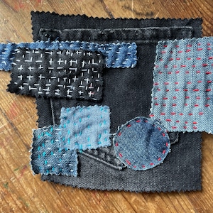 May include: A patchwork of denim fabric scraps sewn together with different coloured thread. The fabric is a mix of blue and black denim, with some pieces having a white or red thread. The fabric is arranged in a random pattern, with some pieces overlapping.