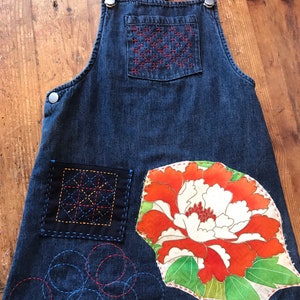 Puede incluir: Un vestido de peto de mezclilla azul oscuro con varios diseños bordados. El vestido tiene un bolsillo grande en la parte delantera con un patrón floral rojo y blanco. El bolsillo está rodeado de costuras negras. El vestido también tiene un bolsillo más pequeño en la parte delantera con un patrón geométrico en hilo negro, amarillo y rojo. El vestido tiene una variedad de otros diseños bordados, incluyendo círculos y cuadrados.
