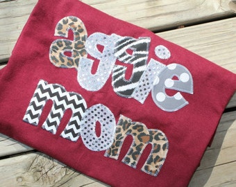 May include: A maroon t-shirt with the words "Aggie Mom" appliqued on the front. The letters are made of various patterned fabrics, including leopard print, zebra stripes, and polka dots. The shirt is folded on a wooden surface.